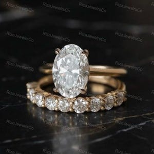 May include: A rose gold engagement ring set featuring a large oval-cut centre stone held by four prongs, paired with a matching band encrusted with a row of round-cut stones. The metal has a warm, polished finish, and the stones are clear and faceted.
