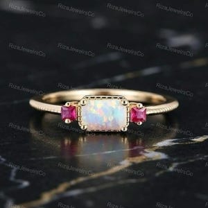 May include: A gold ring featuring a central rectangular opal with a multi-colored iridescent play of color, flanked by two small square-cut red rubies. The band has a thin, smooth profile with a delicate textured detail near the stone setting.