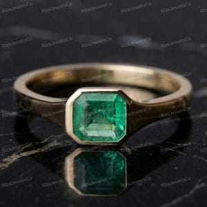 May include: A gold ring featuring a single emerald-cut green emerald gemstone set in a bezel setting. The band is smooth and polished, showcasing a clean design that highlights the vibrant green color of the central stone and the metallic luster.