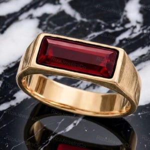 Puede incluir: Un anillo de oro con una piedra preciosa larga, rectangular y de color rojo intenso engastada horizontalmente en el centro. La banda es lisa y pulida, con una superficie superior plana que enmarca la piedra alargada. El diseño es limpio y simétrico.