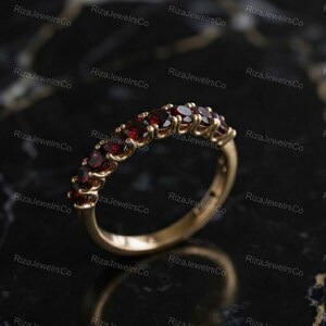Könnte beinhalten: Ein Goldring mit einer Reihe von neun rund geschliffenen, tiefroten Granat-Edelsteinen in einer gemeinsamen Krappenfassung. Der Ring hat eine polierte Metalloberfläche und ein geschwungenes Profil, das die leuchtend roten Steine in einer Reihe zeigt.