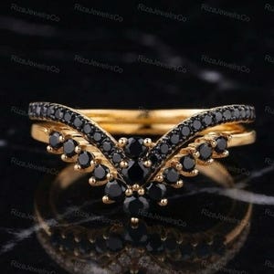 May include: A gold V-shaped ring featuring a row of small, round, black stones set along the band. The center of the chevron design is accented with a cluster of larger round black stones, creating a pointed, symmetrical appearance.
