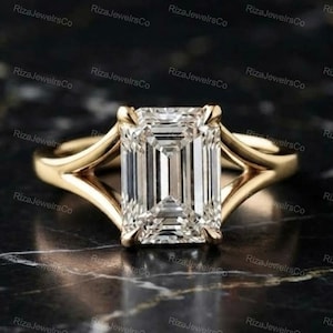 May include: A gold ring featuring a large, rectangular emerald-cut clear gemstone set in a four-prong setting. The band has a split-shank design that tapers as it connects to the central stone, creating a sleek and polished appearance.