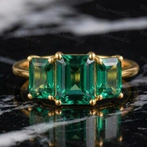 May include: A gold ring featuring three emerald-cut green gemstones arranged in a row. The central stone is larger than the two flanking stones, all held in place by gold prongs on a polished gold band that reflects on the surface below.