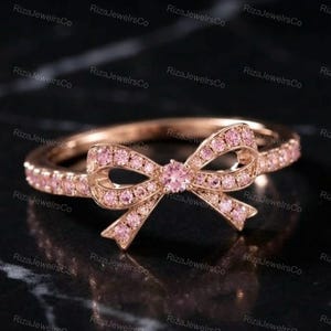 Puede incluir: Un anillo de oro rosa que presenta un diseño central en forma de lazo adornado con pequeñas piedras rosas. La banda también está engastada con una fila continua de piedras rosas a juego, creando una apariencia coherente y detallada en la joya.