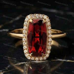 May include: A gold ring featuring a large, rectangular, cushion-cut deep red garnet gemstone at the center. The garnet is surrounded by a halo of small, round, clear white diamonds set in gold. The ring band is thin, smooth, and polished gold.