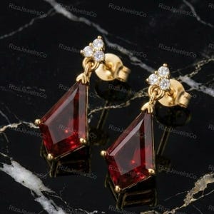 May include: A pair of gold stud earrings featuring a kite-shaped deep red gemstone drop suspended from a small cluster of three clear round stones. The earrings have a polished gold finish and secure with a standard post and butterfly back mechanism.