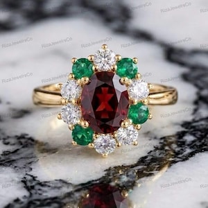 Könnte beinhalten: Ein Goldring mit einem zentralen, oval geschliffenen, tiefroten Granat, umgeben von einem Kranz aus abwechselnd rund geschliffenen grünen Smaragden und klaren weißen Diamanten. Die Ringschiene ist aus poliertem Gold und blütenförmig gefasst.