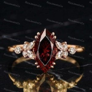 Op de afbeelding: Een roségouden ring met een grote, dieprode granaat in marquise-slijpvorm als middensteen, geflankeerd door twee clusters van kleine, helderwitte accentstenen in marquise- en ronde slijpvorm op een dunne, gepolijste roségouden ring.
