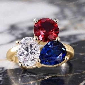 Könnte beinhalten: Ein Goldring mit drei markanten Edelsteinen: einem runden roten Stein, einem ovalen klaren weißen Stein und einem birnenförmigen blauen Stein. Die Steine sind in einem polierten Goldring in einer Cluster-Anordnung fest miteinander verbunden.