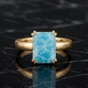 May include: A gold ring featuring a rectangular, light blue Larimar gemstone with a marbled pattern. The stone is held in place by four gold prongs on a smooth, polished gold band. The ring is centered and reflects on the surface below.