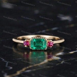 May include: A gold ring featuring a central emerald-cut green gemstone flanked by two smaller square-cut red gemstones. The band has a textured, ribbed design along the sides, and the stones are set in a prong setting on a thin metal band.