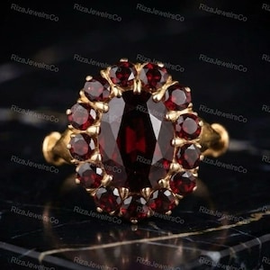 Puede incluir: Un anillo de oro con una gran piedra preciosa granate de color rojo intenso y corte ovalado en el centro, rodeada por un halo de catorce piedras granates más pequeñas de corte redondo. La banda es de oro amarillo y el engaste sujeta las piedras.