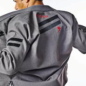 Pode incluir: Uniforme cinza de artes marciais com listras pretas no ombro e na manga. A palavra "TEAM" é impressa em vermelho nas costas, junto com um raio vermelho. O uniforme é feito de um tecido texturizado.