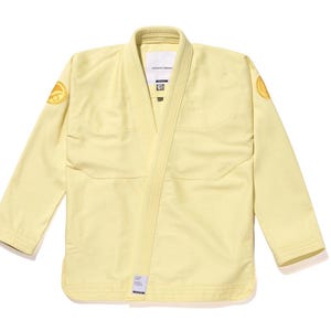 Op de afbeelding: Een lichtgele vechtsportuniform, of gi, met een wikkelontwerp. De gi heeft een kraag, zakken en patches op de schouders. De mouwen en randen hebben een contrasterende gebroken witte bies. Het uniform is gemaakt van een duurzame stof.