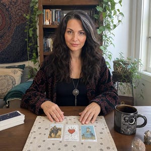K&ouml;nnte beinhalten: Eine Person mit langem dunklem Haar mit grauen Str&auml;hnen, bekleidet mit einem schwarzen Oberteil und einer dunkel gemusterten Strickjacke, sitzt an einem Holztisch mit drei Tarotkarten auf einem beigen Tuch, einem Kartendeck und einer schwarzen Tasse mit der Aufschrift The Moon.
