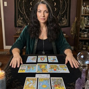 K&ouml;nnte beinhalten: Ein dunkelgr&uuml;ner Samtmantel &uuml;ber einem schwarzen Oberteil, dazu eine silberne Halskette mit Halbmond- und Sternanh&auml;nger. Auf einem schwarzen Tuch liegen zw&ouml;lf Tarotkarten in einer Auslage, erg&auml;nzt durch eine Kristallkugel und zwei Amethystspitzen.