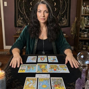May include: A dark green velvet robe worn over a black top, featuring a silver crescent moon and star pendant necklace. A black cloth displays twelve tarot cards arranged in a spread, accompanied by a clear crystal ball and two amethyst points.