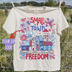 May include: Cream-colored t-shirt with a colorful graphic design. The design features the words "SMALL TOWN FREEDOM" with illustrations of buildings, flowers, and a water tower. The zip code "90782" is also included.