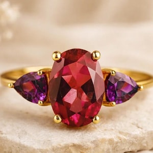 Puede incluir: Un anillo de oro con una gran gema roja de talla ovalada flanqueada por dos gemas moradas en forma de pera. El anillo se exhibe sobre una superficie de piedra clara, resaltando el diseño y el color de la joya.
