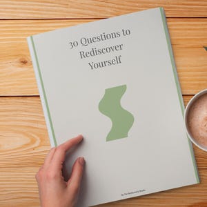 May include: A book titled "30 Questions to Rediscover Yourself" with a green abstract design, next to a cup of coffee on a wooden surface. The book is being touched by a hand.