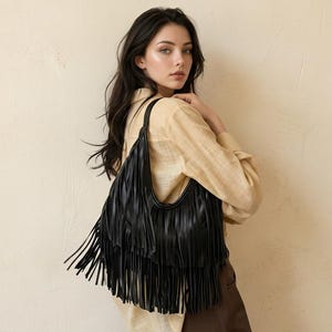 Peut inclure: Un sac à bandoulière noir de style hobo orné de plusieurs couches de longues franges pendantes sur le devant. Le sac présente une ouverture supérieure incurvée et une bandoulière unique, fabriqué dans un matériau sombre et lisse.
