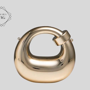 Puede incluir: Un bolso dorado con forma ovalada y redondeada, con un asa grande y circular. El bolso tiene una superficie lisa y reflectante y un pequeño cierre rectangular donde se conecta el asa. El texto "LUNARA BAG" está en la esquina superior izquierda.