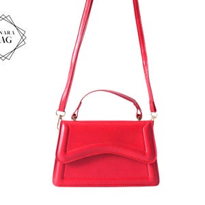 May include: A vibrant red handbag with a top handle and a long, adjustable shoulder strap. The bag is rectangular with a curved flap closure. The brand name "LUNARA BAG" is displayed in a geometric design in the upper left corner.