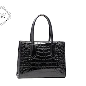 Peut inclure: Un sac à main noir brillant avec une texture en peau de crocodile. Le sac a deux poignées et une forme rectangulaire. Le nom de la marque "LUNARA BAG" est affiché dans un logo géométrique.