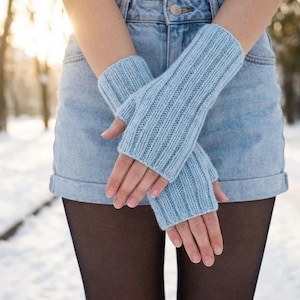May include: Light blue knitted fingerless gloves. The gloves are ribbed and cover the hands up to the knuckles. The person is wearing a denim skirt and black tights. The background is a snowy outdoor scene.