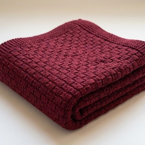 May include: Folded burgundy blanket with a woven texture. The blanket is made of a soft material and has a decorative border. The blanket is folded neatly, showing the texture and color. The blanket is a cozy home textile.