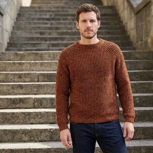 May include: A man wearing a brown knit sweater and dark blue jeans stands on a stone staircase. The sweater has a ribbed texture and a crew neck. The background features a stone staircase.