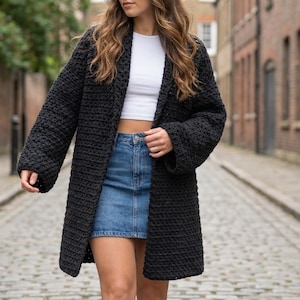 May include: A long, black, open-front cardigan with a textured, chunky knit design. The cardigan is worn over a white crop top and a denim skirt. The sleeves are long and the cardigan falls to mid-thigh. The background is a cobblestone street.
