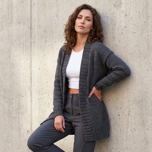 May include: A woman wearing a dark gray, open-front cardigan sweater over a white top and gray pants. The cardigan has long sleeves and a ribbed texture. The woman is leaning against a concrete wall.