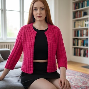 May include: A bright pink, textured, cropped jacket with three-quarter sleeves. The jacket is worn over a black top and paired with a black skirt. The outfit is worn while sitting on a gray sofa.