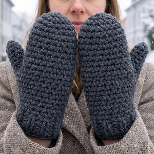 May include: A pair of dark gray, hand-crocheted mittens. The mittens are made of a thick yarn, with a textured stitch pattern. The mittens are shown against a snowy outdoor background, suggesting winter wear.