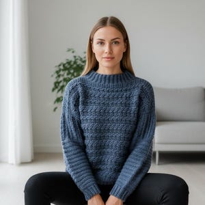 May include: A blue knit sweater with a textured pattern. The sweater has a high neckline and long sleeves. The person is wearing black pants. The background includes a window, a plant, and a gray sofa.