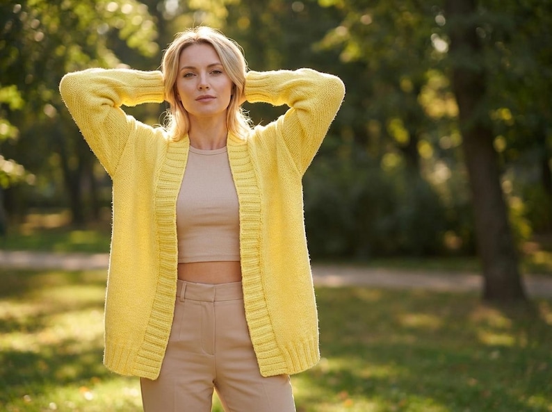 Puede incluir: Una mujer con un c&aacute;rdigan amarillo, un top corto beige y pantalones beige. El c&aacute;rdigan est&aacute; abierto, revelando el top y los pantalones. El fondo es un parque con &aacute;rboles y sol.