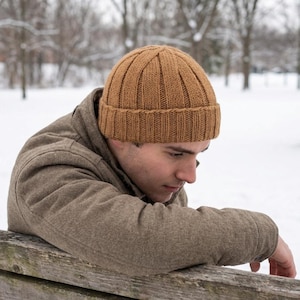 Puede incluir: Un gorro de punto acanalado marrón. El gorro lo lleva una persona que lleva una chaqueta marrón, apoyada en un banco de madera. El fondo es un parque nevado con árboles desnudos.