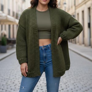 May include: An olive green, open-front cardigan with long sleeves. The chunky knit sweater is paired with a matching olive green top and blue jeans. The cardigan has two front pockets and a relaxed fit.