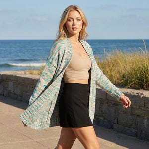 Puede incluir: Una mujer con un c&aacute;rdigan estampado en azul claro, turquesa y blanco, un top corto beige y una minifalda negra. Camina por un sendero de hormig&oacute;n cerca del mar, con la playa y la hierba de fondo.