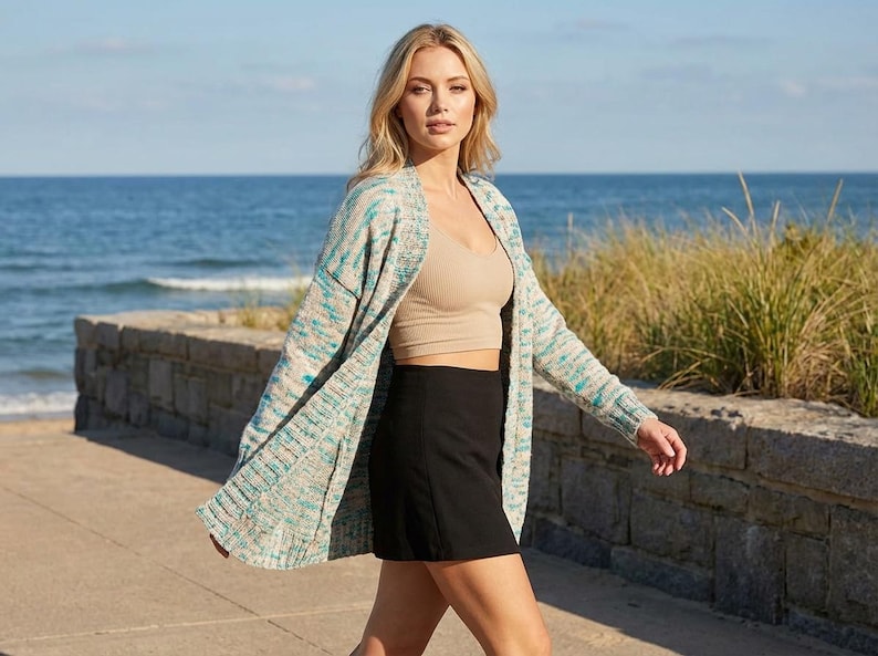 Puede incluir: Una mujer con un c&aacute;rdigan estampado en azul claro, turquesa y blanco, un top corto beige y una minifalda negra. Camina por un sendero de hormig&oacute;n cerca del mar, con la playa y la hierba de fondo.
