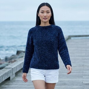 May include: A person wearing a navy blue, long-sleeved sweater with a textured appearance. They are also wearing white shorts. The background features a body of water and a wooden pier.
