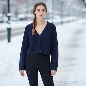 May include: A woman wearing a navy blue cropped cardigan with a V-neck and button closure, paired with black leggings. The background shows a snowy street scene with blurred trees and buildings.