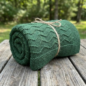 Könnte beinhalten: Gerollte grüne Strickdecke mit Zickzackmuster, mit Bindfaden gebunden. Die Decke besteht aus einem weichen, strukturierten Material und wird auf einer verwitterten Holzoberfläche präsentiert. Die Decke hat die Farbe Waldgrün.