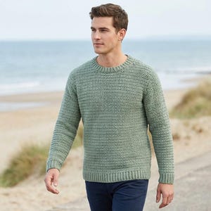 Könnte beinhalten: Ein hellgrüner Pullover mit Rundhalsausschnitt und langen Ärmeln. Der Pullover hat ein strukturiertes Strickmuster und wird mit dunkelblauen Hosen getragen. Der Hintergrund zeigt einen Strand und das Meer.