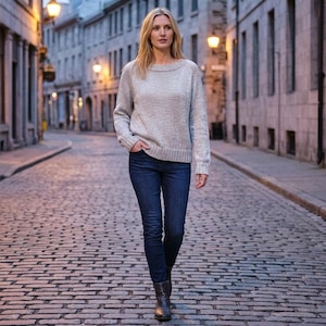 Puede incluir: Una mujer con un suéter gris claro, jeans azul oscuro y botas negras camina por una calle adoquinada. El suéter tiene cuello redondo y mangas largas. Los jeans son ajustados y las botas son de tobillo.