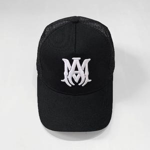 Pode incluir: Boné de beisebol preto com parte traseira em malha e aba curva. O boné apresenta um logotipo branco bordado na frente. O logotipo é um monograma estilizado das letras "A" e "M".