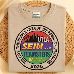 Op de afbeelding: Een beige Gildan heavy cotton t-shirt met een cirkelvormige afbeelding. De afbeelding bevat de tekst "WE GOT THE PEOPLE WE GOT THE POWER UNION STRONG" rond een ontwerp met de tekst "UTLA SEIU TEAMSTERS AALA Local 2010 2026".