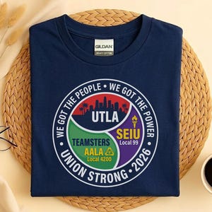 Op de afbeelding: Marineblauw t-shirt met een cirkelvormige afbeelding. De afbeelding bevat de tekst "WE GOT THE PEOPLE", "WE GOT THE POWER", "UTLA", "TEAMSTERS AALA Local 4200" en "SEIU Local 99". Het shirt heeft ook de tekst "UNION STRONG 2026".